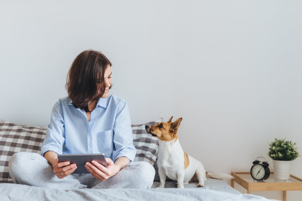 Woman and her dog sitting on a bed next to a nightstand with a clock.