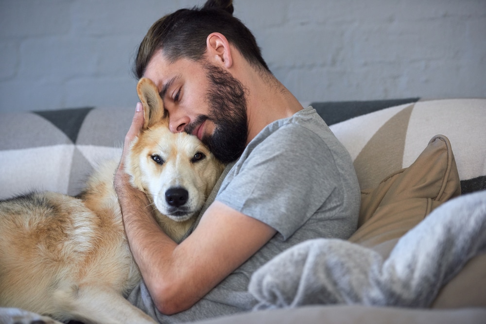 An owner laying on their couch and giving a hug to their dog.