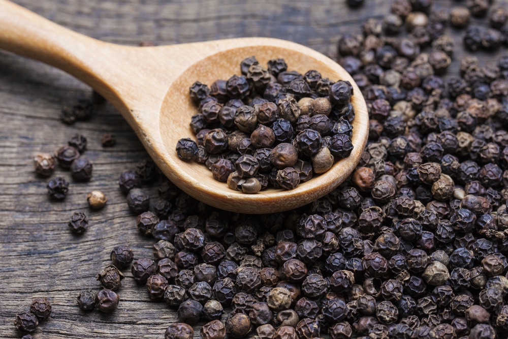 Black pepper scattered all around a wooden spoon with some in it as well.
