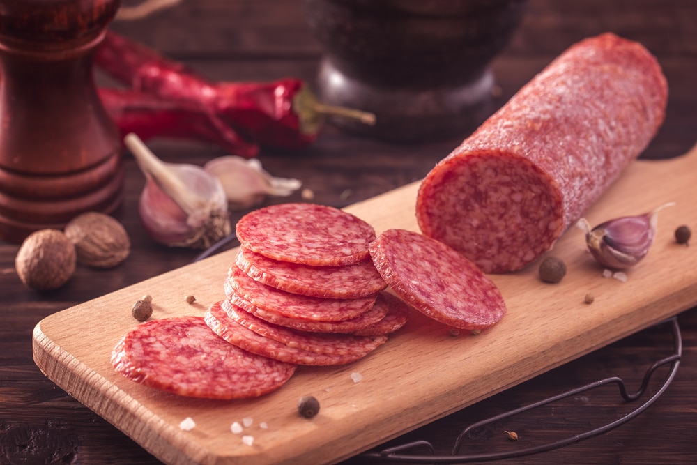 Some salami and slices on a cutting board with other food in the background.